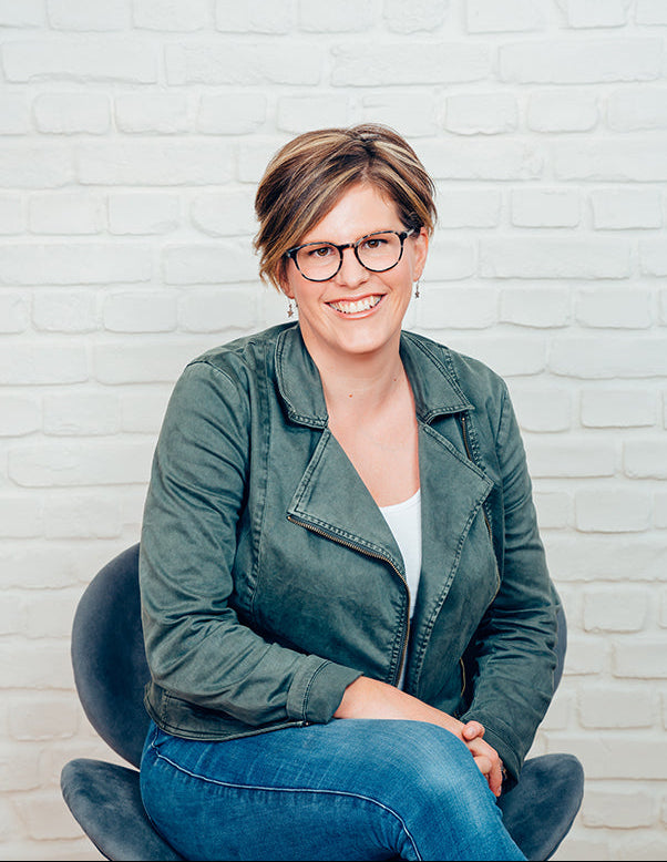 A smiling woman with short brown hair and glasses sits in a dark chair against a white painted brick wall. She is wearing an olive green jacket over a white top and blue jeans.